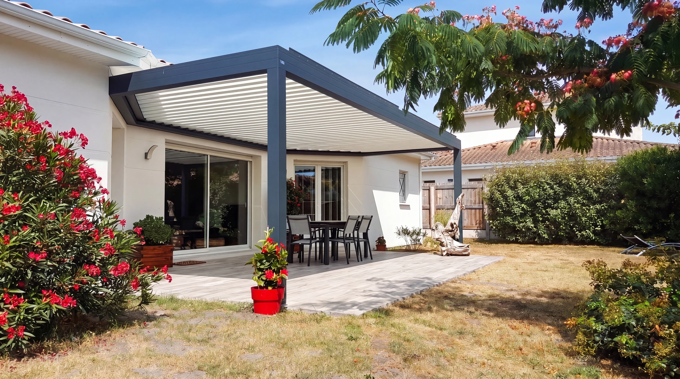 Terrasse extérieure avec pergola bioclimatique à lames orientables en aluminium anthracite, fixée en applique sur façade de maison plain-pied blanche
